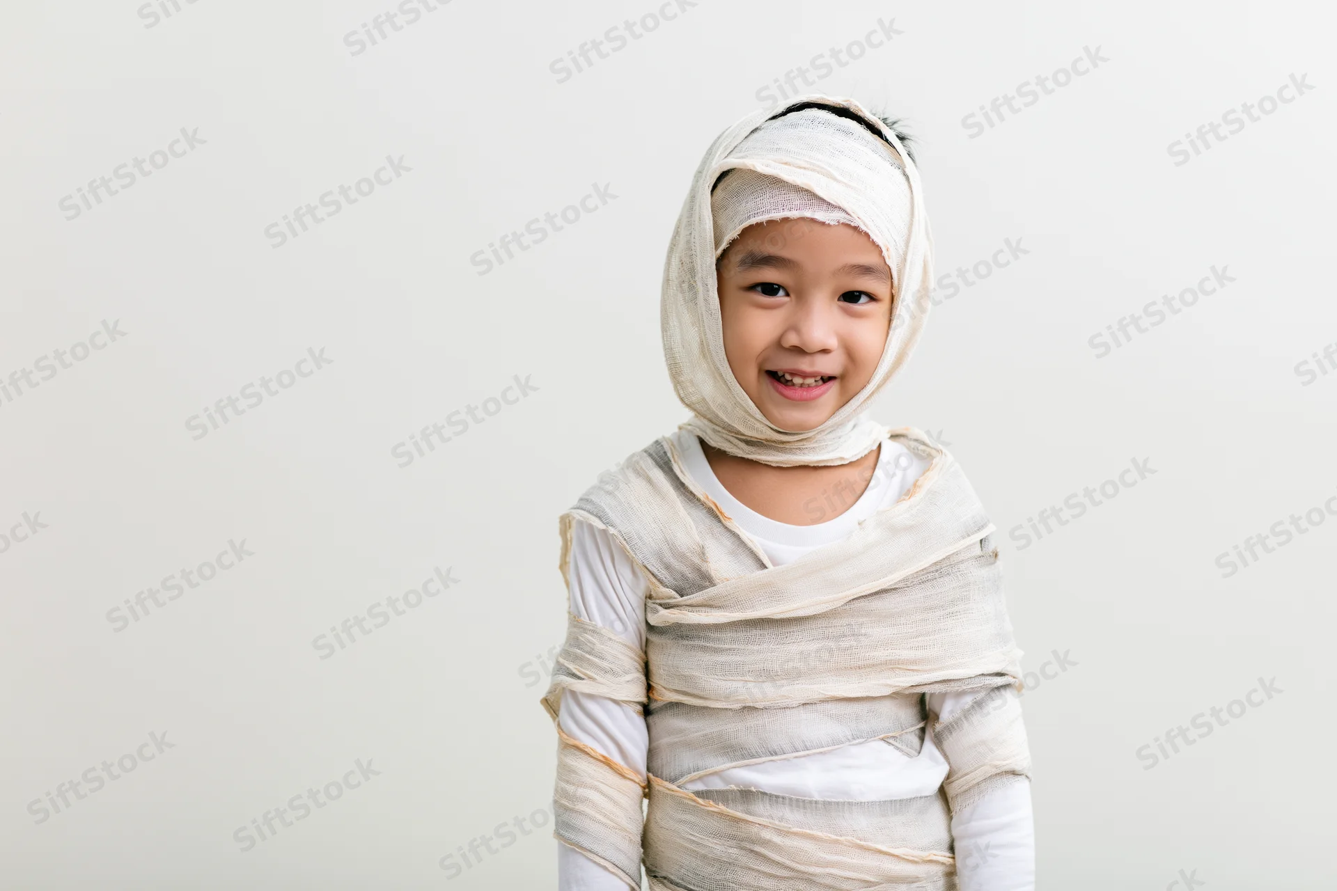 Young asian boy wrapped in white bandages for a homemade mummy halloween costume, smiling at camera against plain background