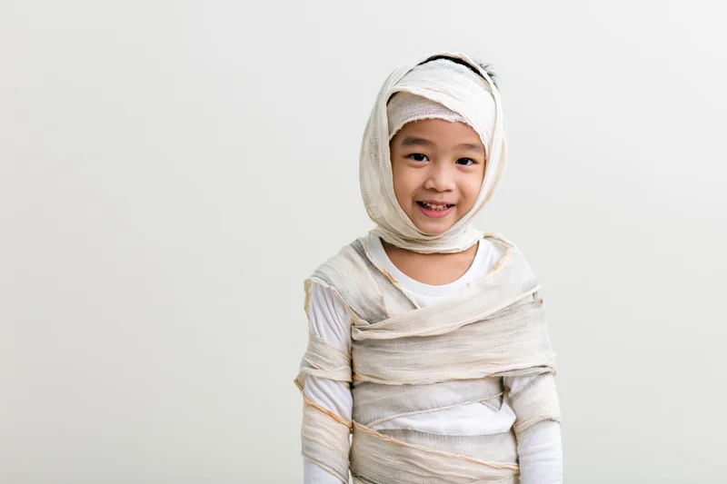 Young asian boy wrapped in white bandages for a homemade mummy halloween costume, smiling at camera against plain background