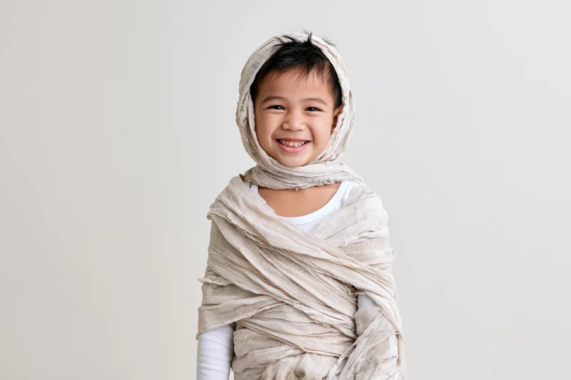 Young asian boy wrapped in beige fabric as a homemade mummy costume smiling in studio