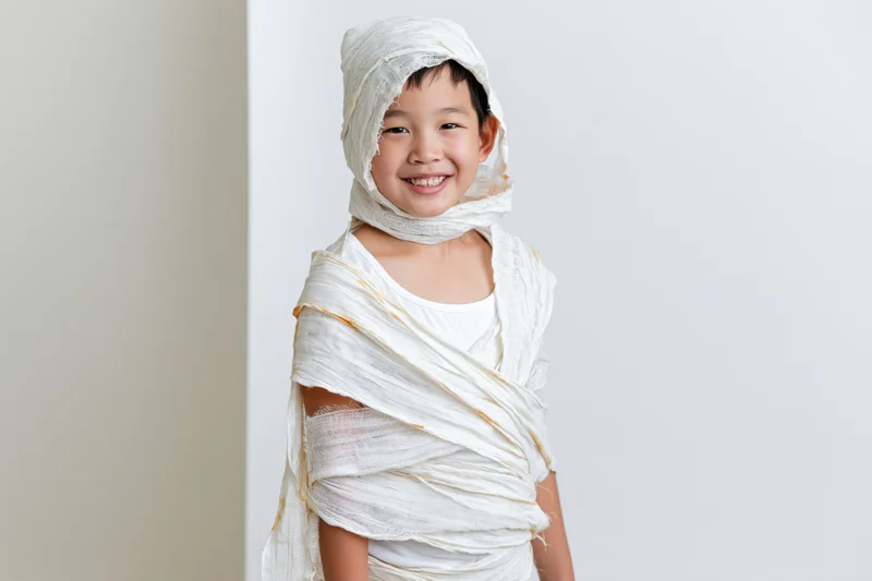 Young asian boy smiling while wrapped in white fabric as a homemade mummy costume for halloween