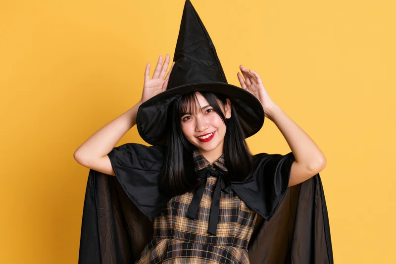 Young asian woman in witch costume smiling playfully with hands on hat against yellow background