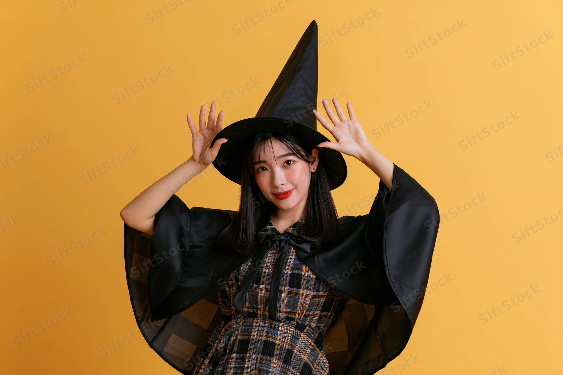 Young asian woman in witch costume posing playfully with hands raised against orange background