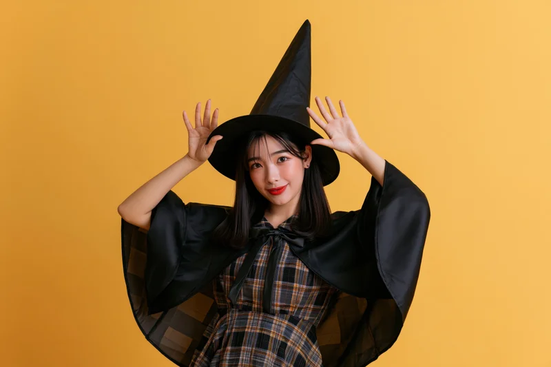 Young asian woman in witch costume posing playfully with hands raised against orange background