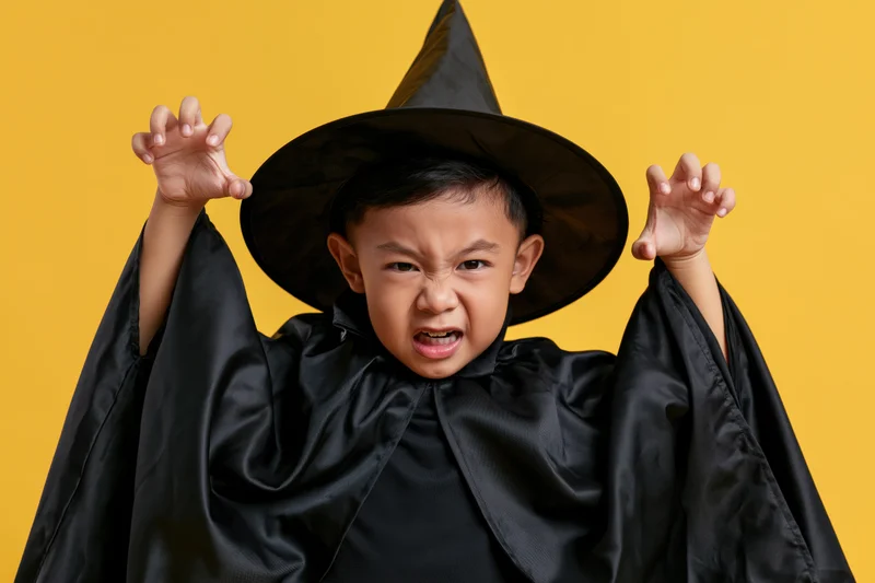 Young southeast asian boy in black wizard costume posing with raised hands against yellow background