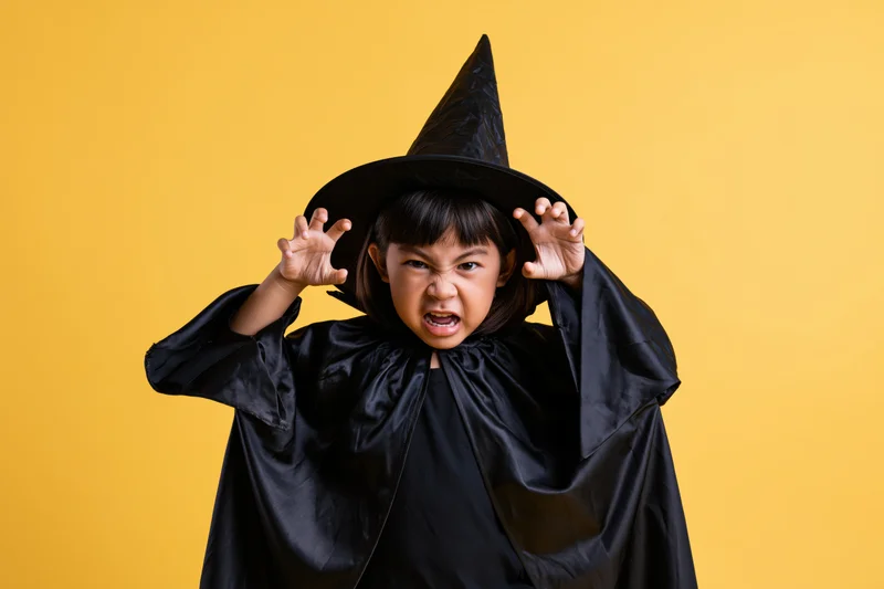Young southeast asian boy in black wizard costume making angry face with hands raised against yellow background