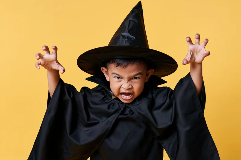 Young southeast asian boy in black wizard costume with arms raised and playful expression against yellow background