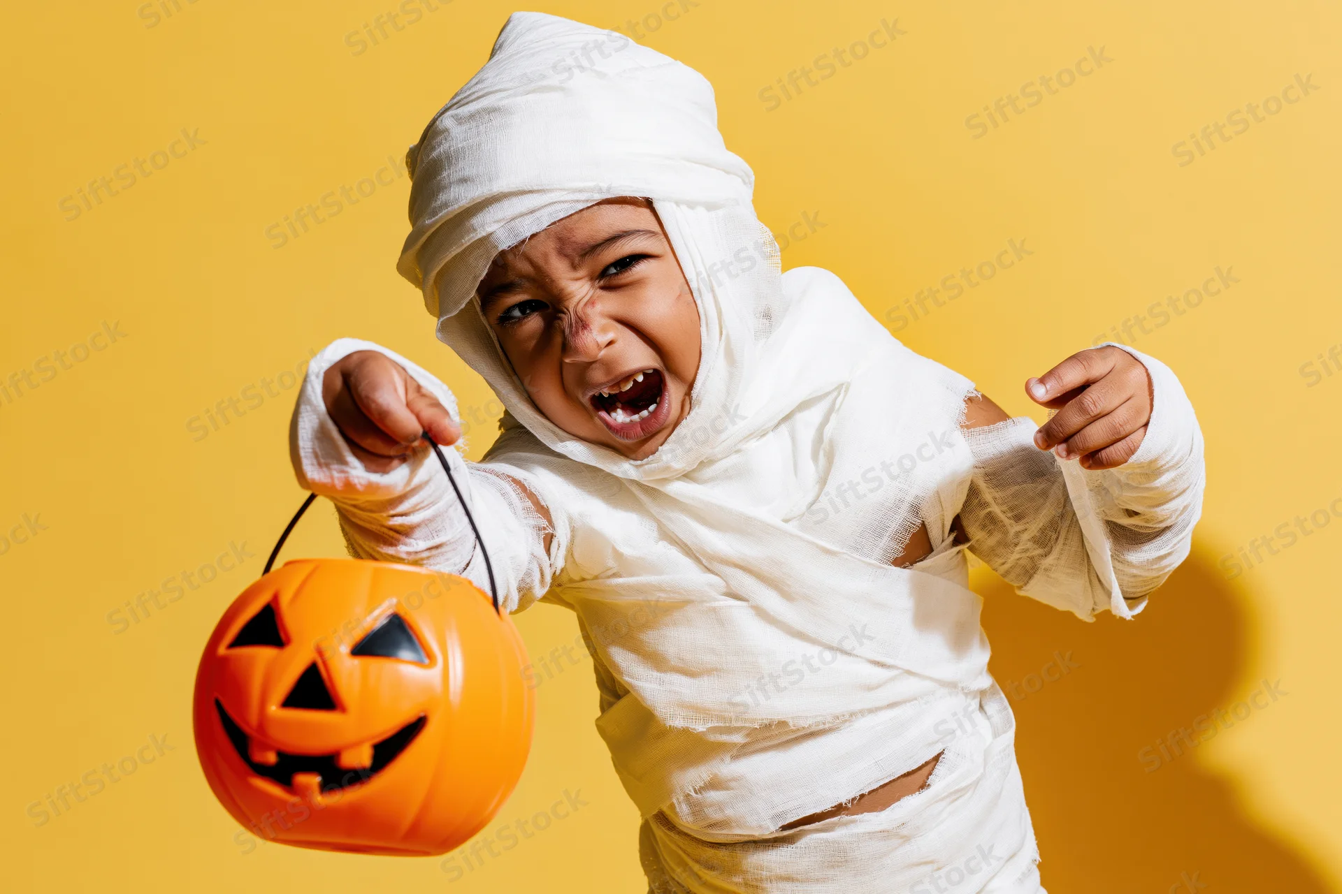 Young child in mummy costume holding orange pumpkin bucket, posing against yellow background