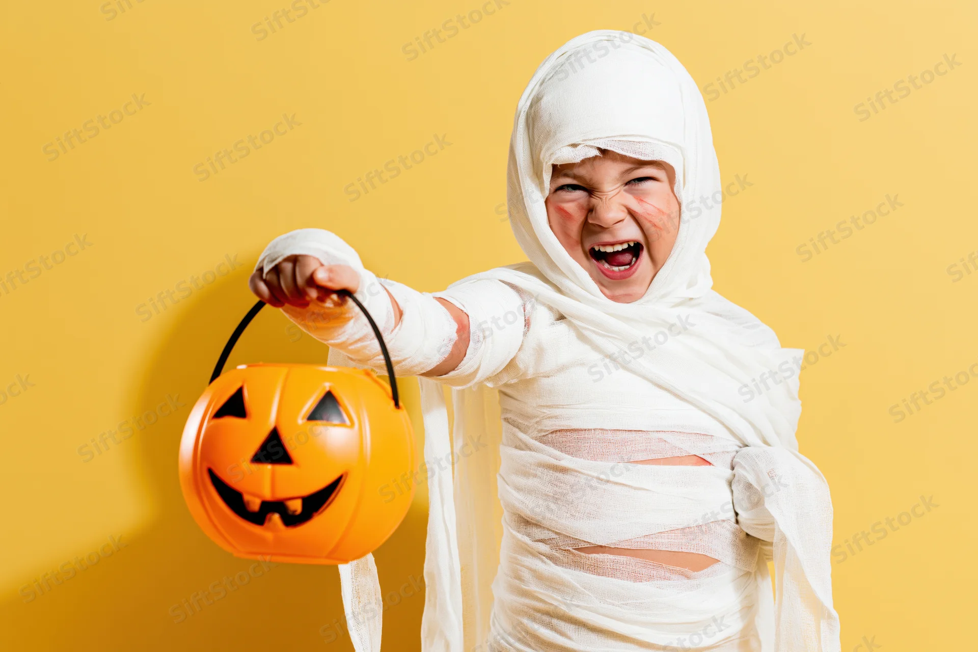Child in mummy costume holding orange pumpkin bucket against yellow background