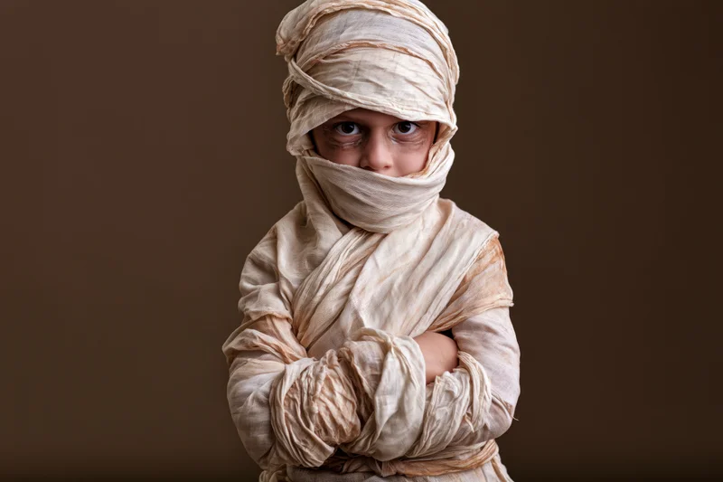 Child wrapped in beige bandages as a mummy costume with arms crossed against brown background