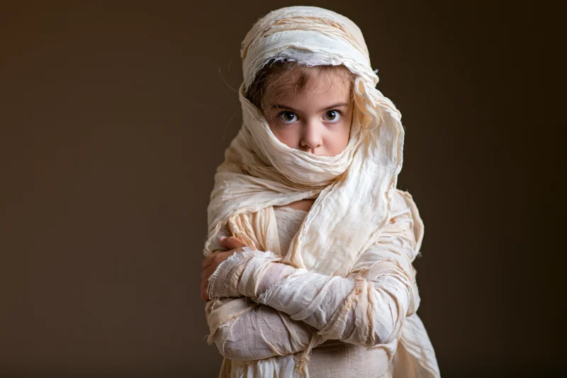 Young girl wrapped in white fabric as a mummy costume for halloween, arms crossed, studio portrait