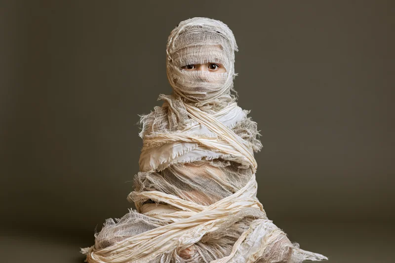 Child wrapped in white bandages as a mummy costume seated against dark background