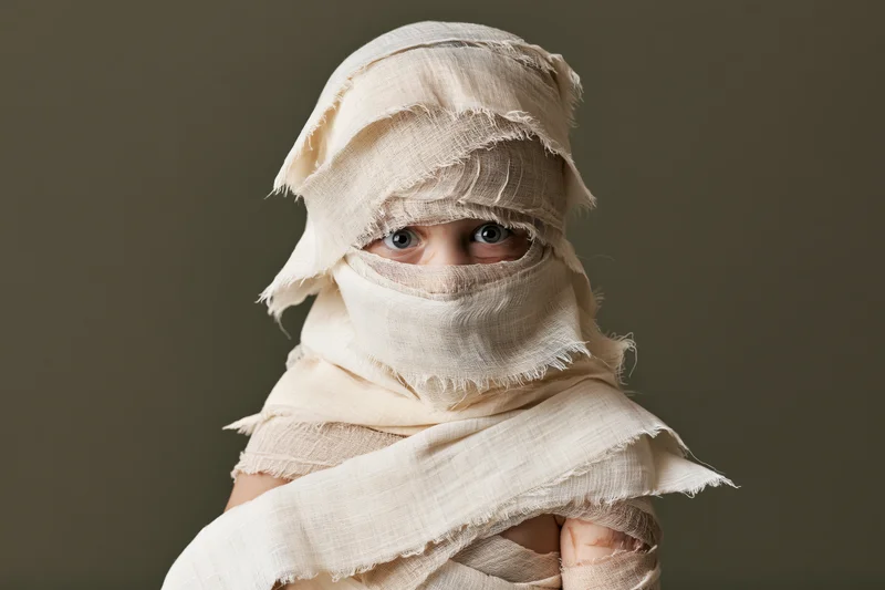 Child wrapped in beige fabric bandages for a mummy costume, eyes visible, studio portrait