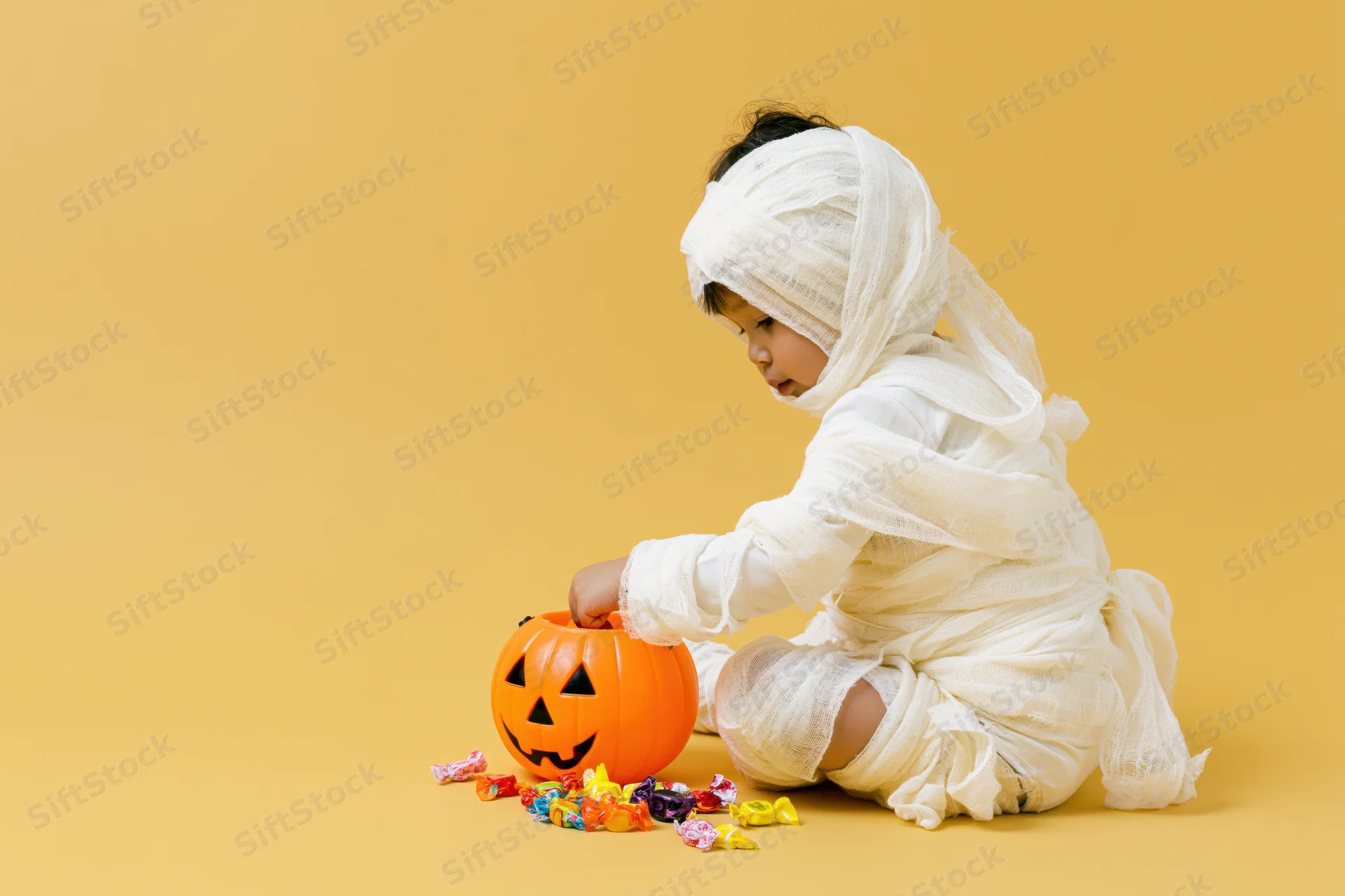 Young child in white gauze mummy costume sitting and sorting candy from orange pumpkin bucket on yellow background