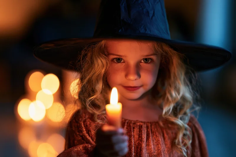 Young girl in witch costume holding lit candle with warm glow and bokeh lights behind