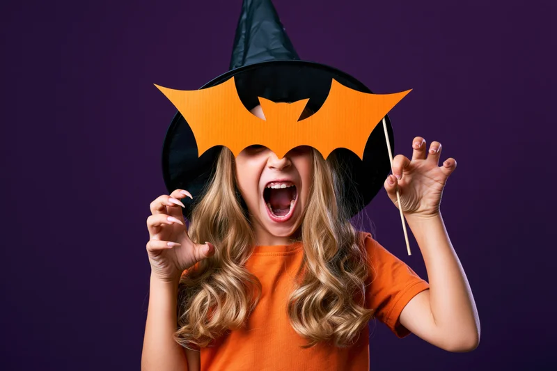 Young girl in witch hat holding orange paper bat mask, shouting playfully against purple background