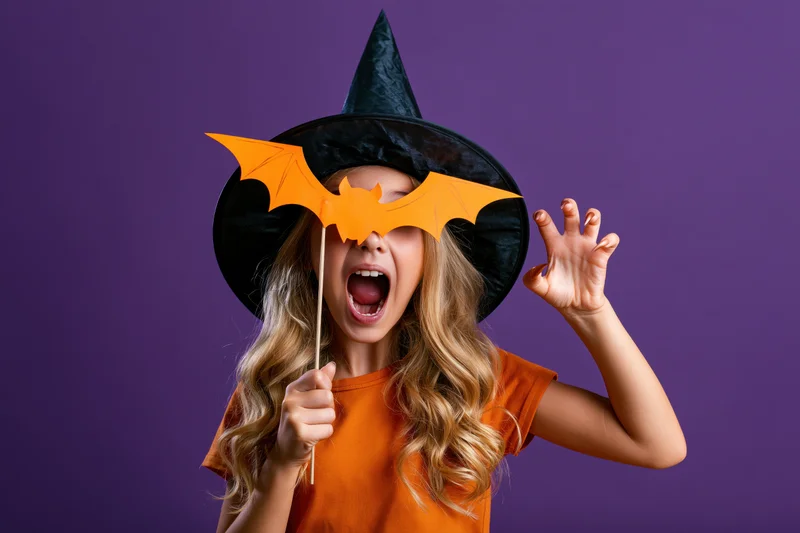 Young girl in witch hat holding paper bat cutout against purple background