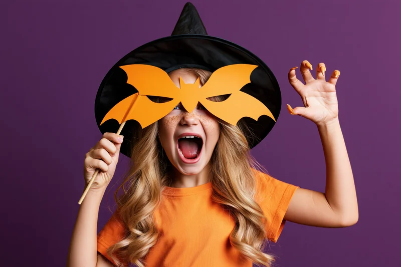Young girl in witch costume holding orange paper bat mask, posing playfully against purple background