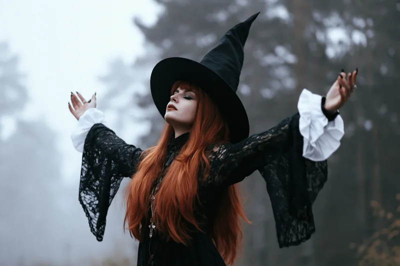 Red-haired woman in black witch costume with arms raised in foggy forest