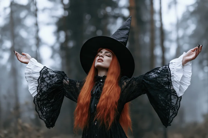 Red-haired woman in black witch costume with arms outstretched in misty forest