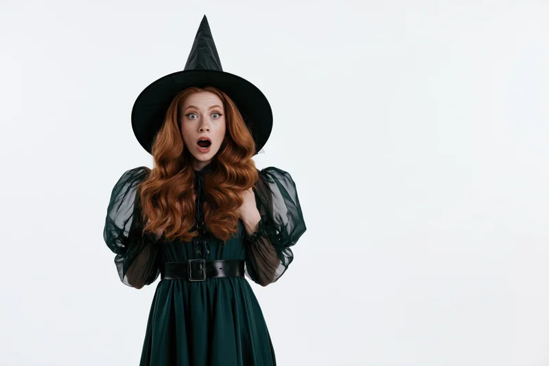 Young redheaded woman in witch costume with surprised expression against white background