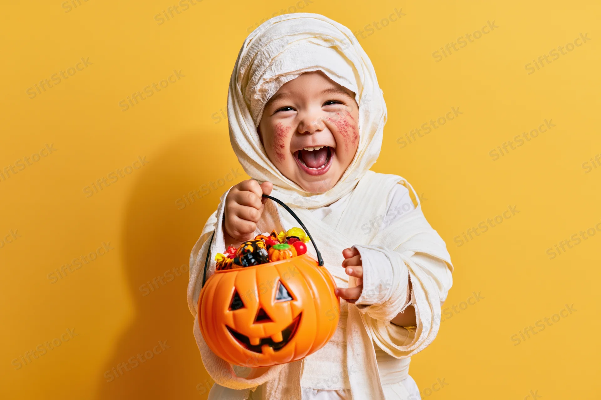 Toddler in white mummy costume holding orange pumpkin bucket filled with candy, smiling against yellow background