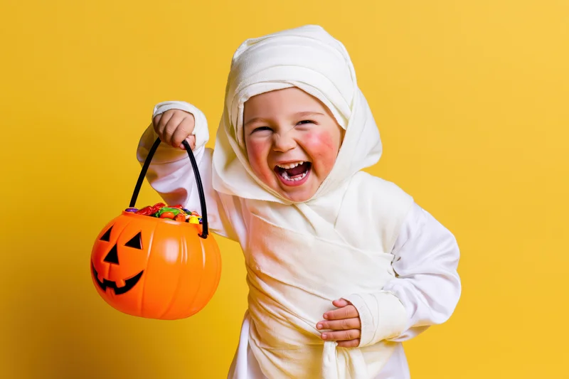 Toddler in white mummy costume laughing while holding orange pumpkin candy bucket against yellow background