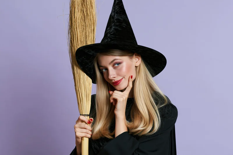 Young woman in witch costume holding broom and posing with hand on chin against purple background