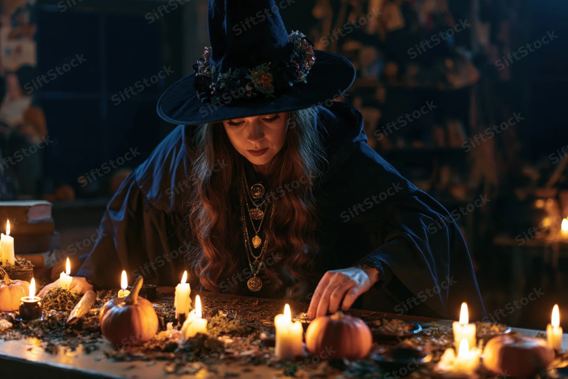 Young woman in witch costume leaning over wooden table with candles and pumpkins during halloween ritual