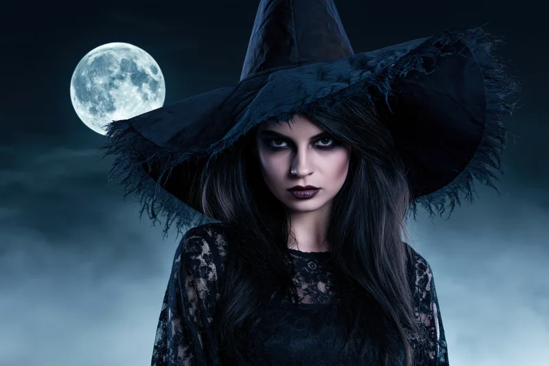 Woman in witch costume with wide-brimmed hat gazing directly at camera under full moon at night
