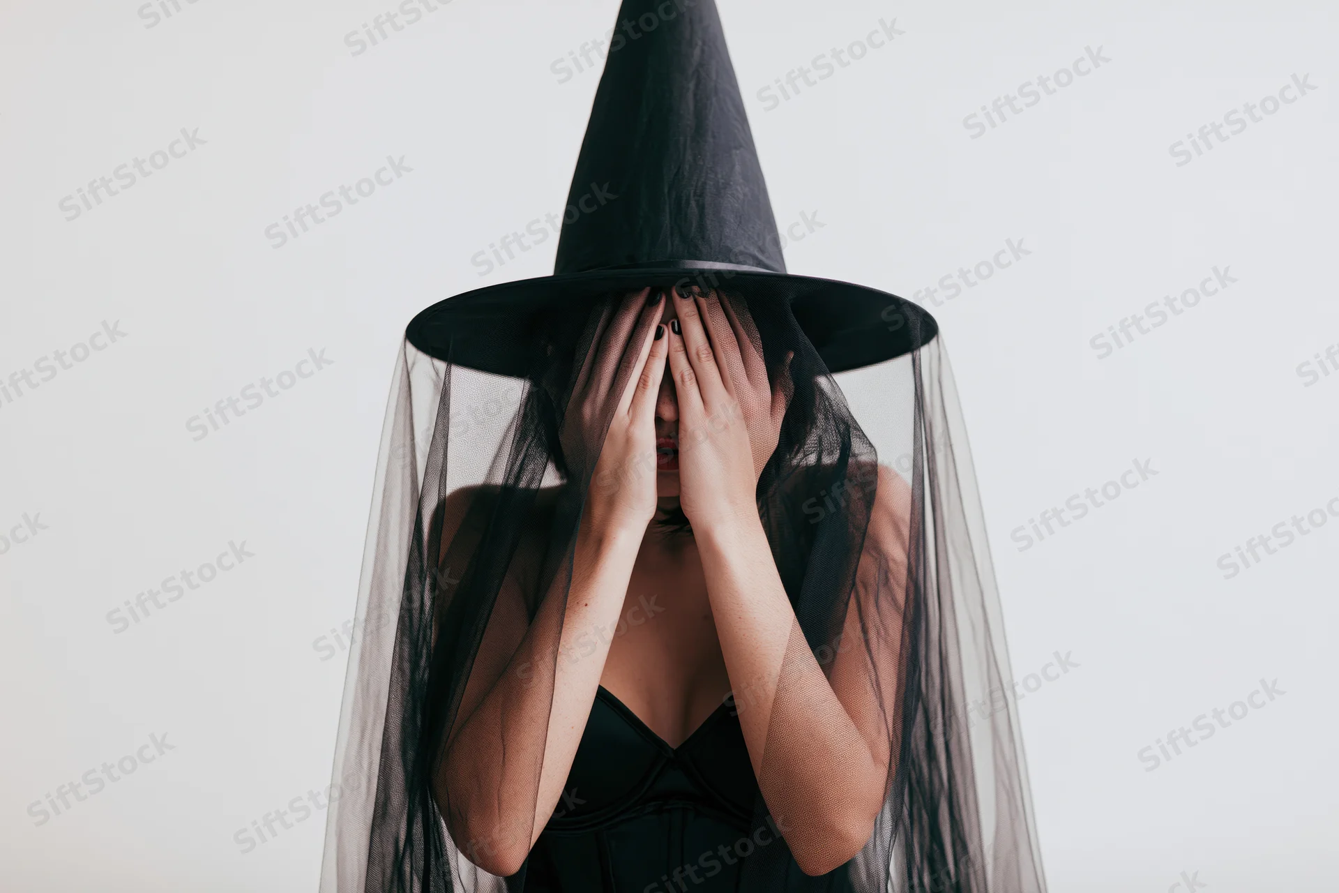 Woman in witch costume covering face with hands, studio portrait