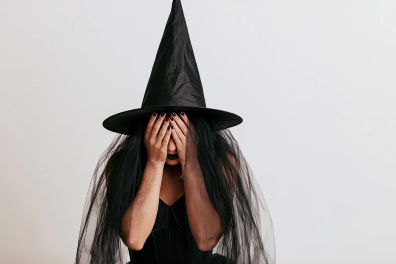 Woman in witch costume covering face with hands against white background