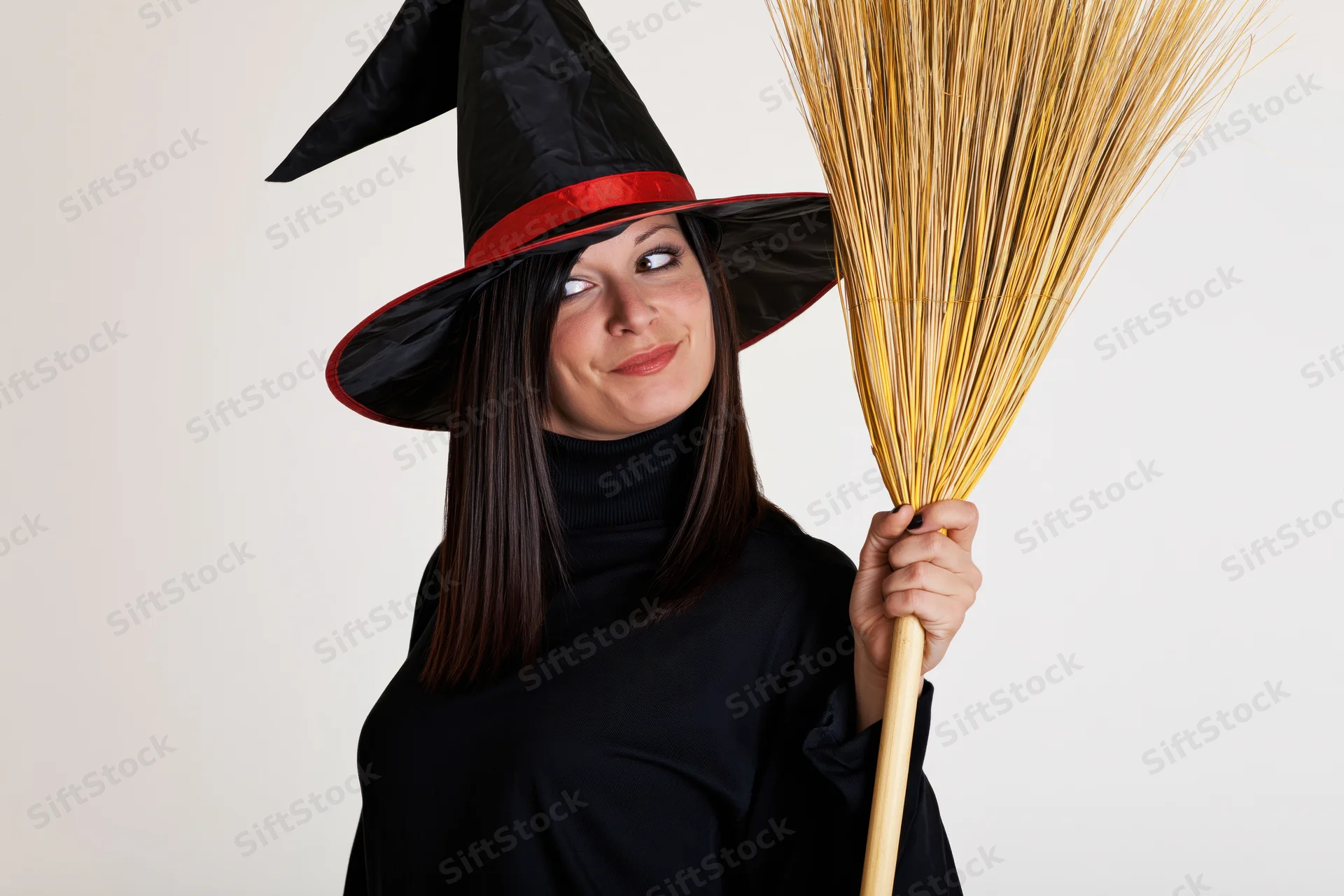 Young woman in witch costume holding straw broom, smiling and looking aside in studio setting