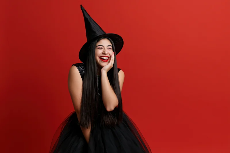 Woman in witch costume laughing with hand on cheek against red background