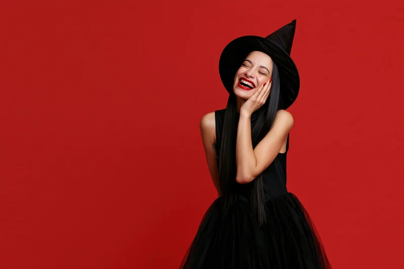Woman in witch costume laughing with hand on face against red background