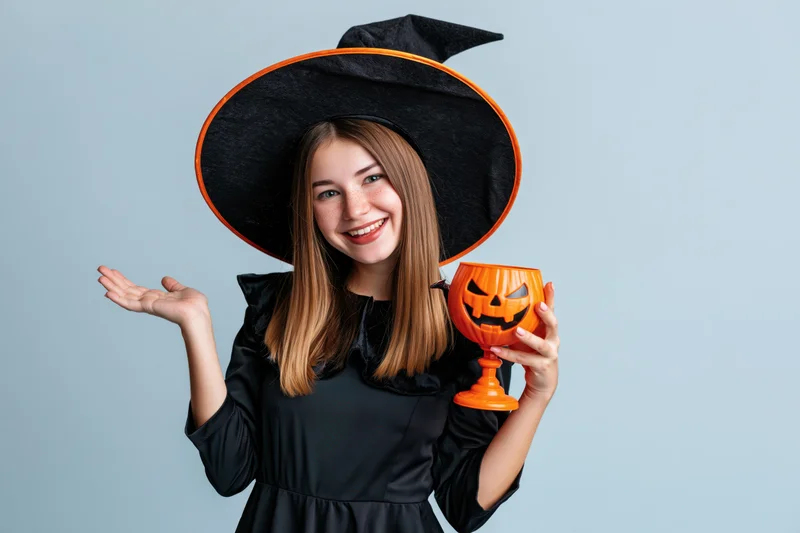 Young woman in witch costume smiling and holding pumpkin cup against light blue background