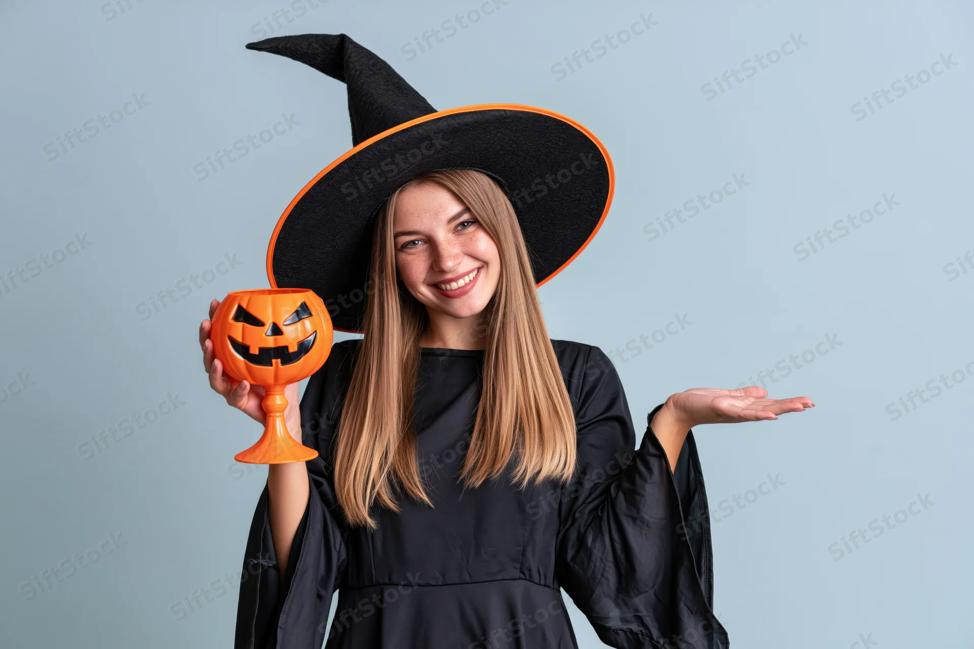 Young woman in witch costume smiling and holding pumpkin cup against light blue background