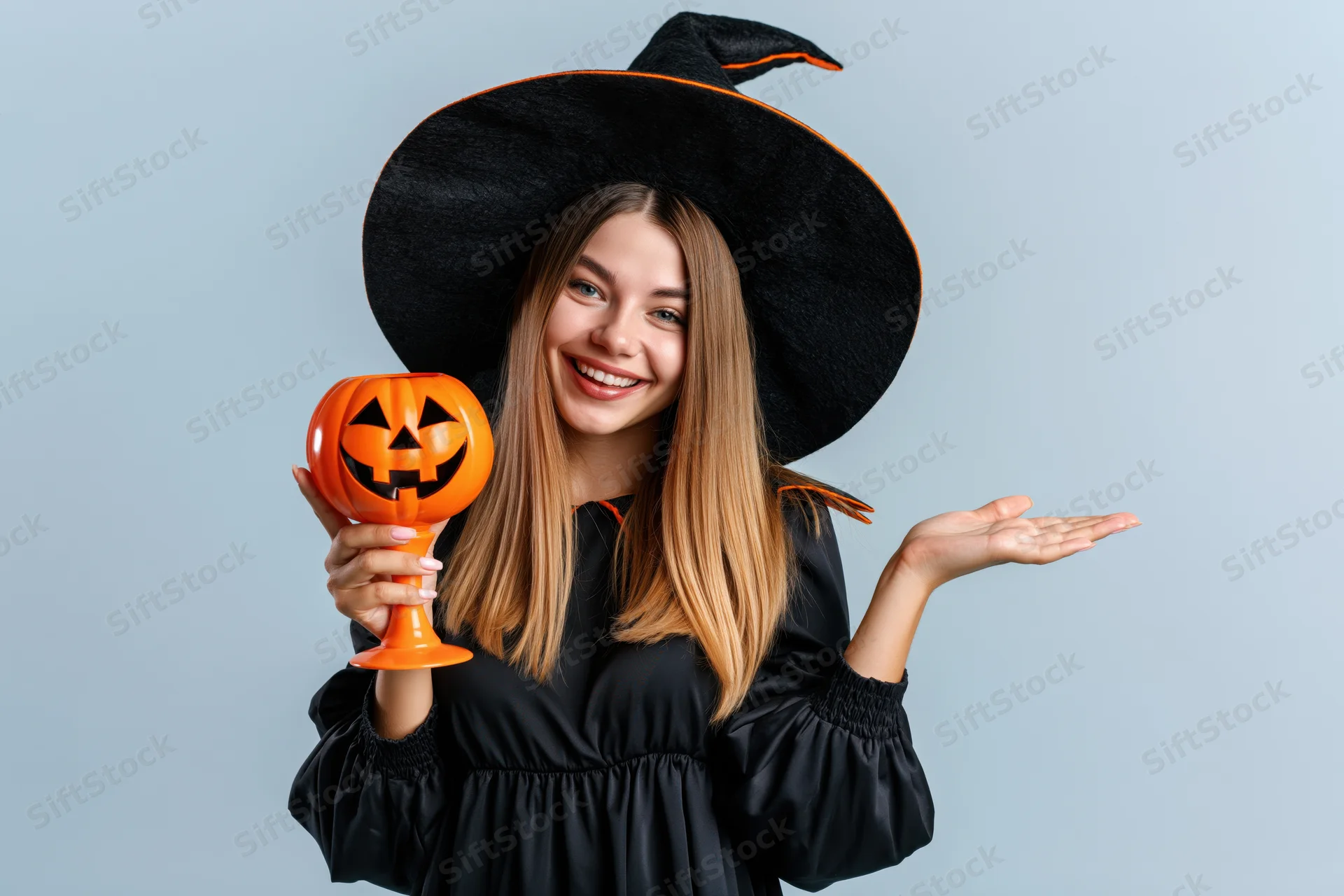 Young woman in witch costume smiling and holding pumpkin-shaped container against light blue background