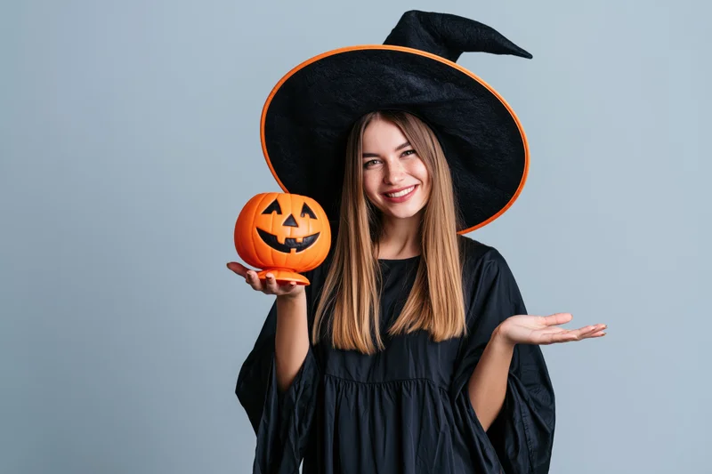 Young woman in witch costume smiling and holding a jack-o-lantern against a plain background