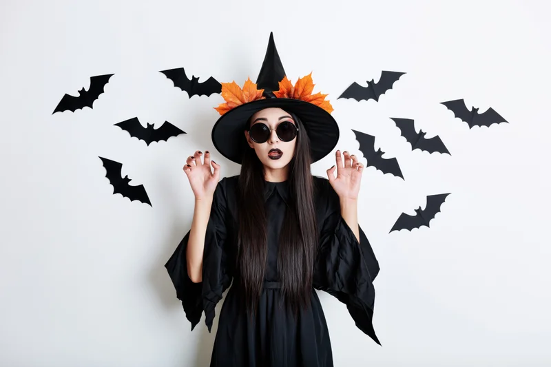 Woman in witch costume with surprised expression surrounded by paper bats on white background
