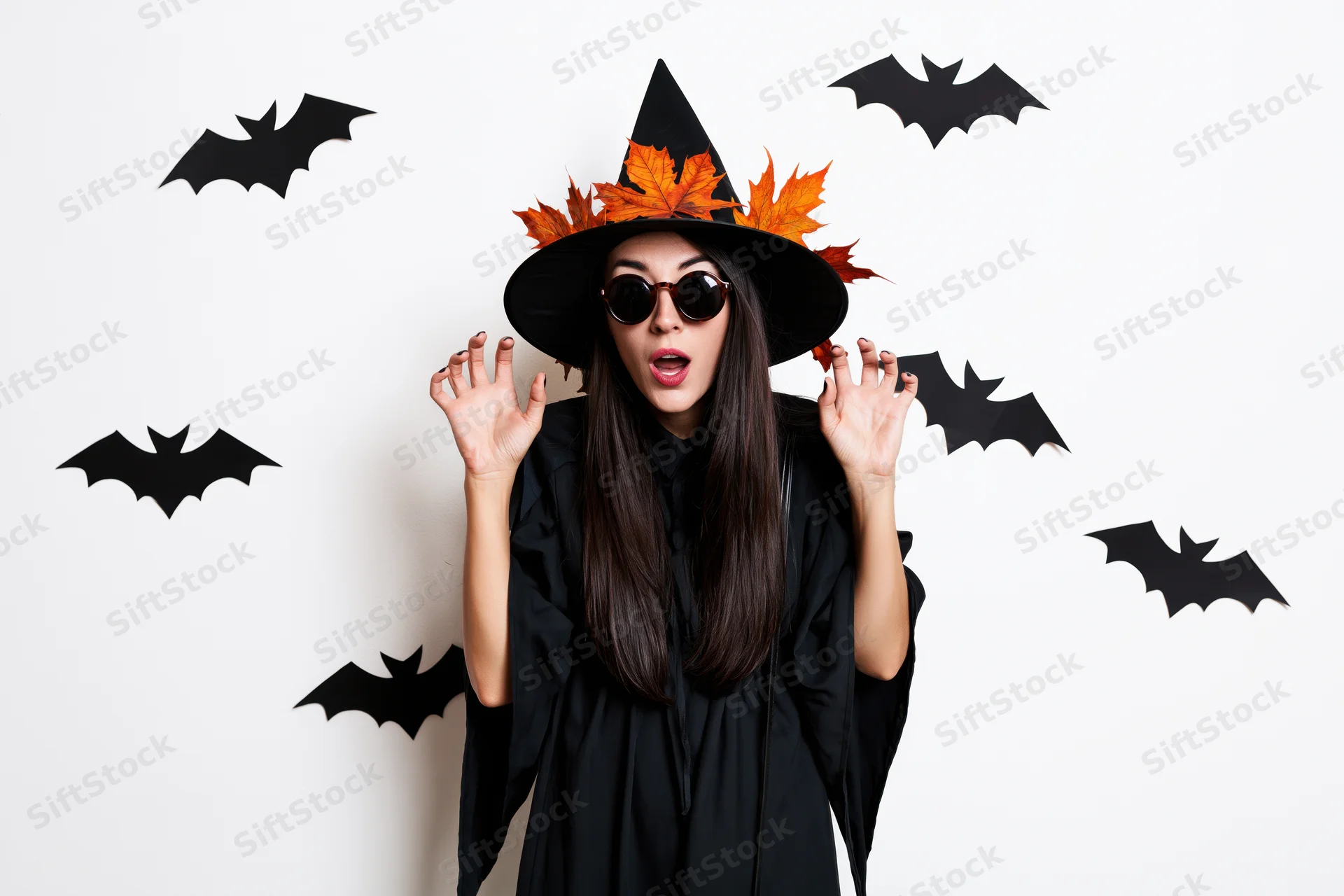 Woman in witch costume with surprised expression wearing sunglasses and hat decorated with autumn leaves, surrounded by paper bats on white background