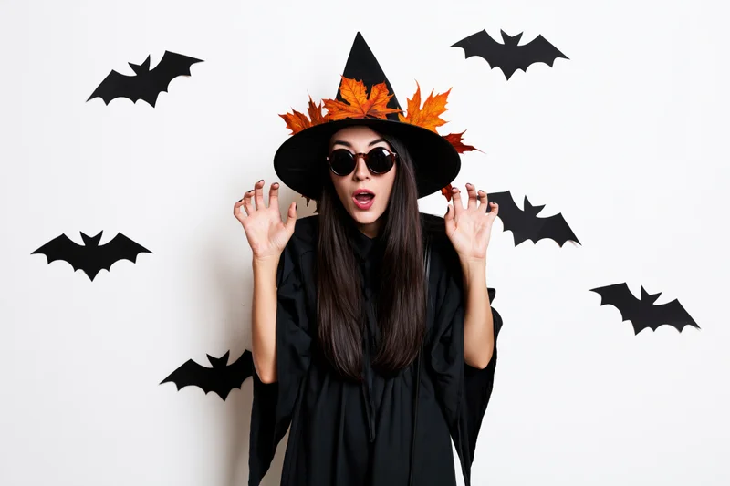 Woman in witch costume with surprised expression wearing sunglasses and hat decorated with autumn leaves, surrounded by paper bats on white background