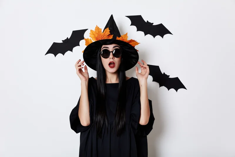 Woman in witch costume with surprised expression and paper bats around her head against white background