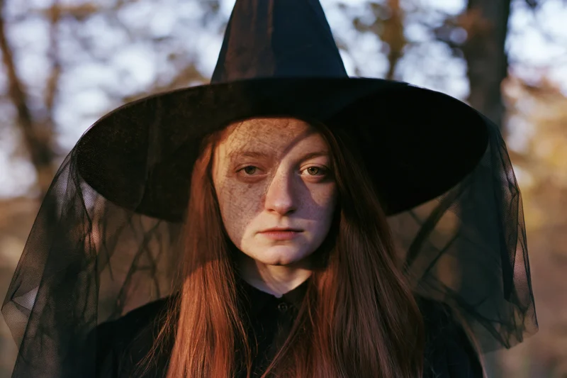 Young woman in witch costume with black pointed hat and sheer veil outdoors in autumn light