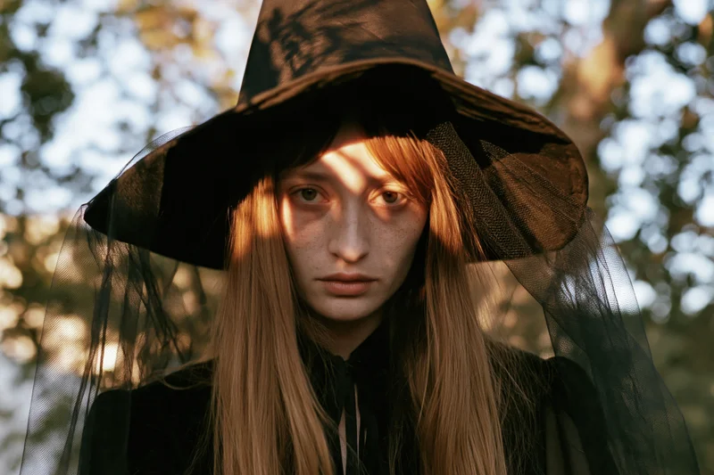 Young woman in witch costume with pointed hat and tulle veil standing in forest at golden hour