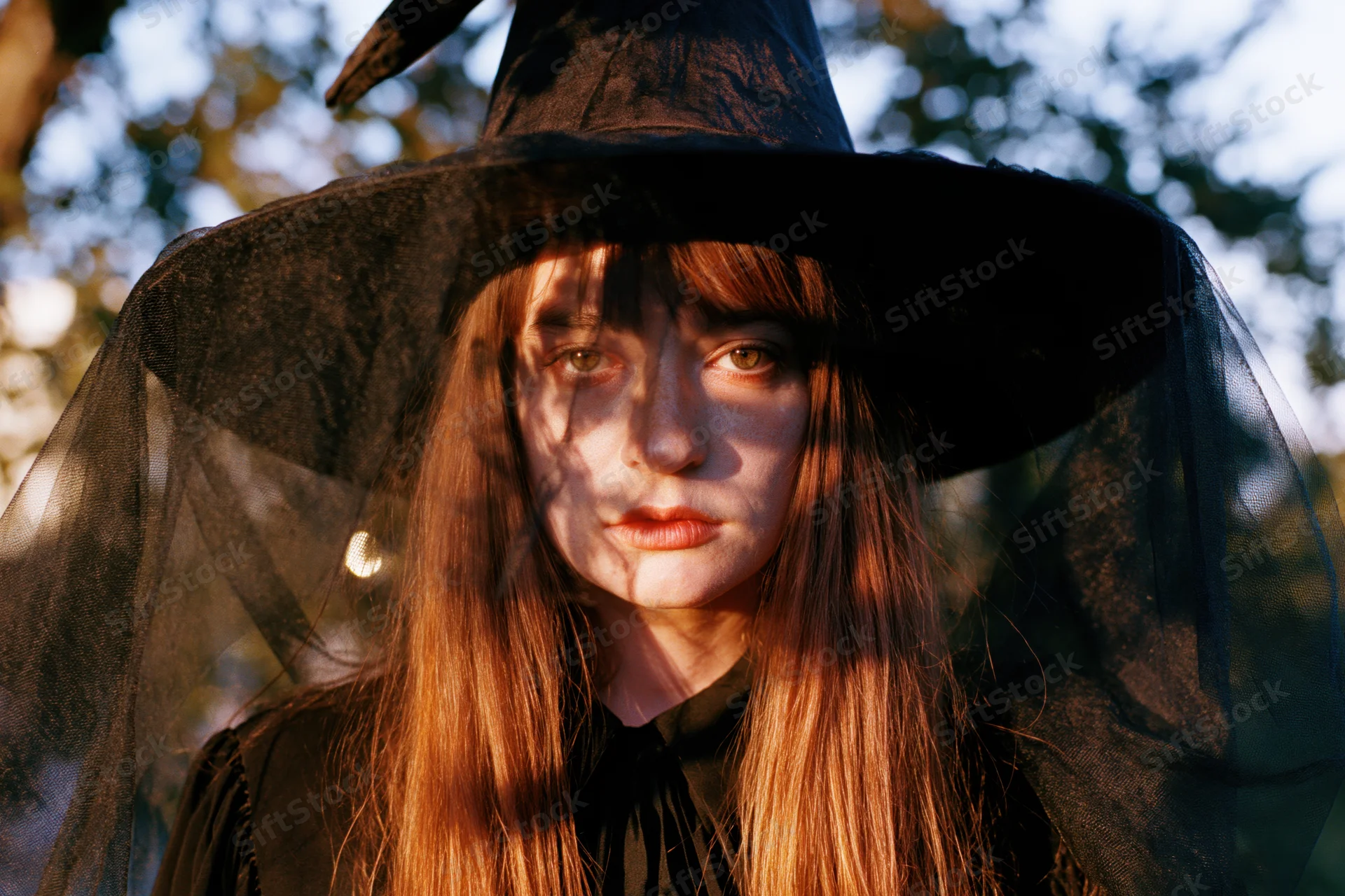 Young woman in witch costume with black pointed hat and sheer veil outdoors in sunlight