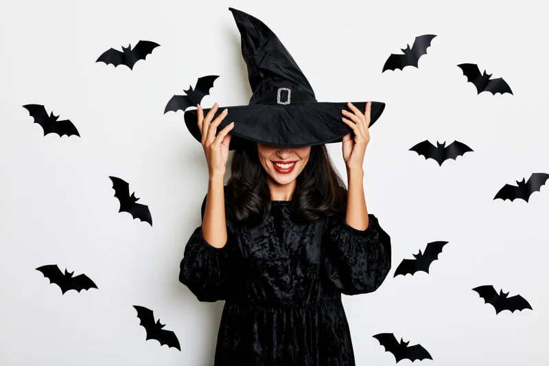 Woman in black witch costume holding hat with bats around her on white background