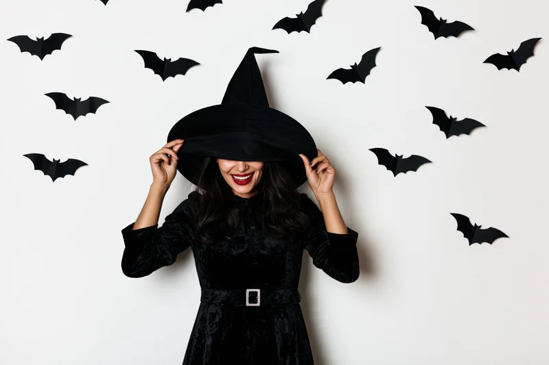 Woman in black witch costume pulling hat over face against wall with paper bats