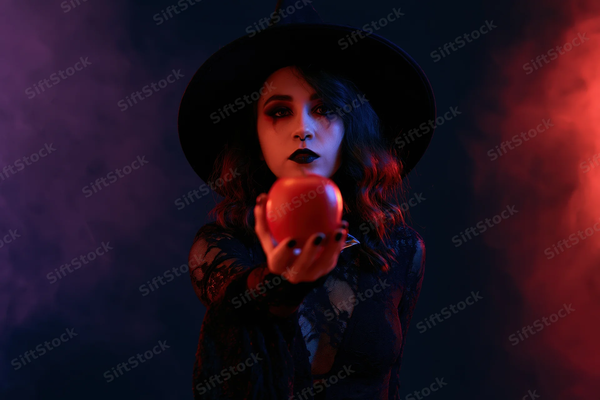 Woman in witch costume holding red apple in smoky studio with purple and red lighting