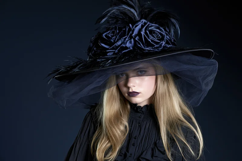 Young woman in wide-brimmed black witch hat with feathers and veil, wearing black gothic blouse, studio portrait