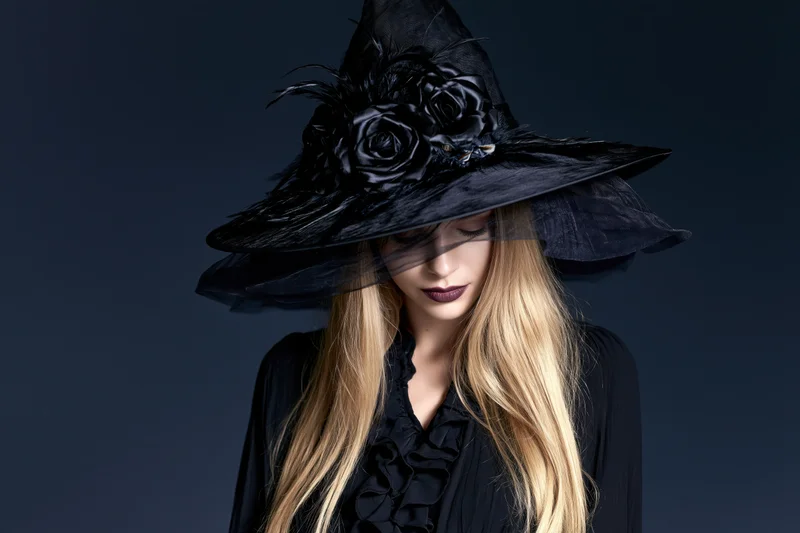 Woman in black witch hat with feathers and roses, wearing dark ruffled dress, looking down against dark background